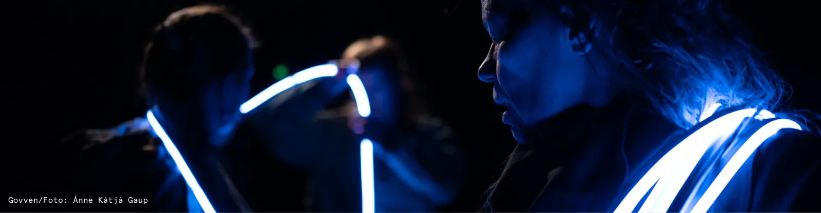 A close up of a woman's face and shoulders seen in profile. It is dark, but you can make up the features of her face. around her neck is coiled a LED light strip that is reminiscent of a lasso. In the distance you can make out two other people, who are also holding the LED light strip.